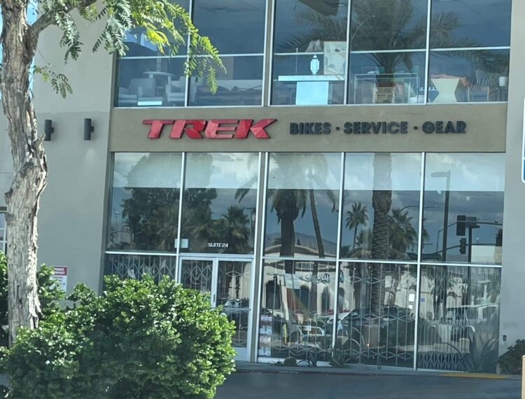 The image shows the front of a TREK bike store with the words "BIKES - SERVICE - GEAR" underneath the main logo. There is also a blue disabled parking sign indicating "PARKING ONLY MINIMUM FINE $250". Reflections of palm trees can be seen in the store's glass windows, and there are some green bushes in the foreground.