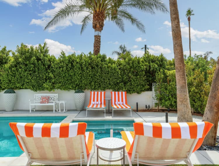 A sunny poolside setting with orange and white striped lounge chairs, tall palm trees, and a lush green hedge in the background.