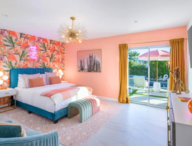 A vibrant bedroom with a blue upholstered bed against a floral wallpaper with flamingos, pink walls, golden curtains, and a view to a garden and pool area through a glass door. The room features a mid-century modern chandelier, a neon sign above the bed, a decorative monkey lamp, and eclectic furnishings.
