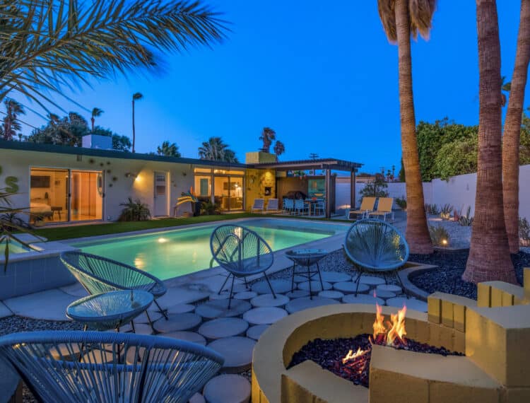 A modern backyard at dusk with a swimming pool and a lit fire pit, surrounded by stylish patio furniture and palm trees, adjacent to a single-story house.