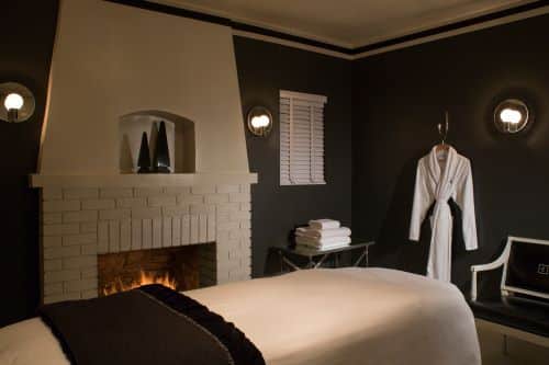 A cozy and elegant room featuring a lit fireplace with a white mantle, a neatly made bed with a dark headboard, flanked by wall-mounted lights, and a bathrobe hanging nearby. The room has dark walls and a contrasting white ceiling, giving off a warm and inviting ambiance.