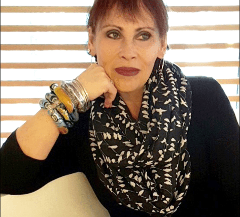 A woman with short red hair wearing dark lipstick and a black top. She has a patterned scarf around her neck and multiple bracelets on her wrist. She's resting her chin on her hand, looking directly at the camera with a neutral expression. Blinds are visible in the background, partially letting light in.