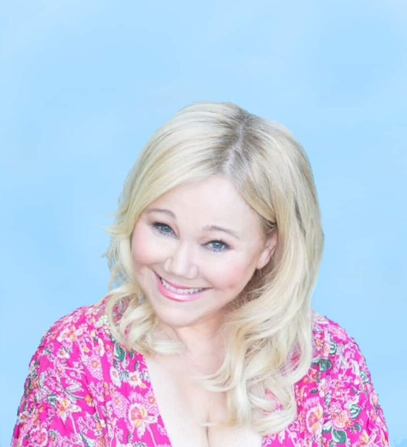 A smiling woman with blonde hair wearing a pink floral top against a soft blue background.