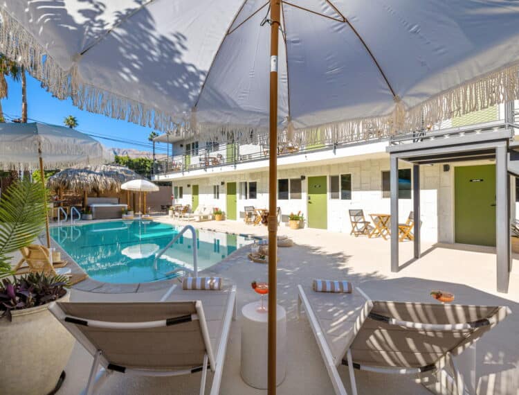 A bright and sunny pool area with lounge chairs and large, fringed umbrellas, surrounded by a white fence and a building with a balcony.