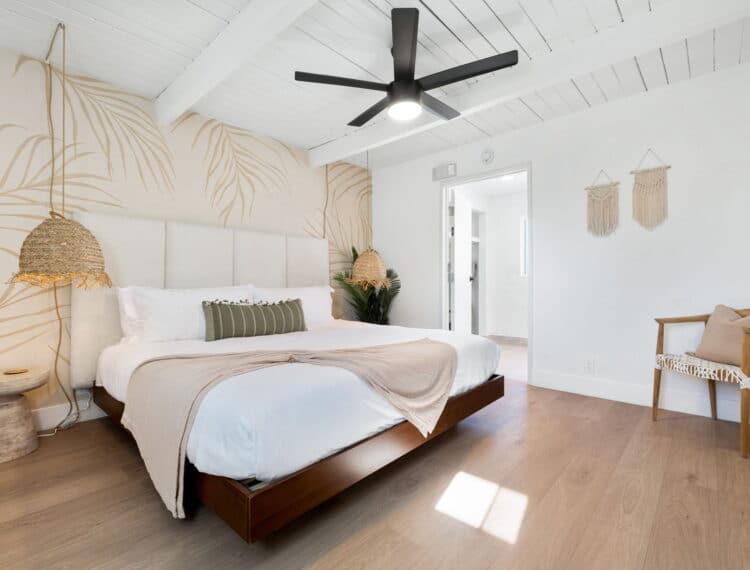 A contemporary bedroom with white walls featuring a large bed with beige and green pillows, a wicker pendant light, wooden flooring, a black ceiling fan, decorative wall weavings, and a chair with a cushion in the corner.