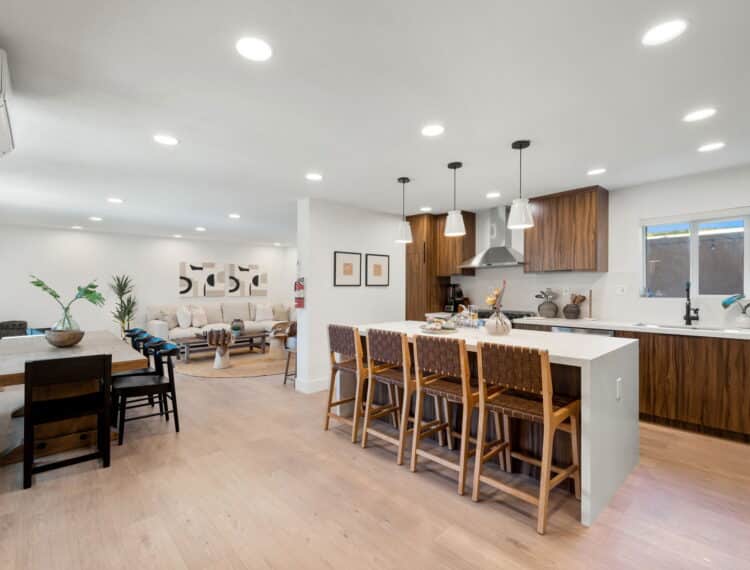 A modern, open-plan kitchen and dining area with a white island and wooden cabinets, complemented by a long wooden dining table with chairs and a living space in the background. The room is well-lit with ceiling spotlights and pendant lights above the island.