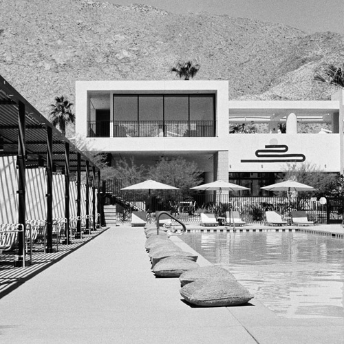 Black and white photo of an outdoor swimming pool with umbrellas and lounge chairs, bean bag loungers on the side, a modern two-story building in the background, and mountains under a clear sky.