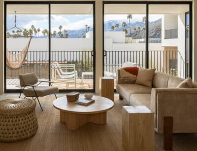 Modern living room with large glass doors opening to a balcony overlooking a mountain landscape, furnished with a beige sofa, wooden coffee tables, wicker chairs, and a hammock.
