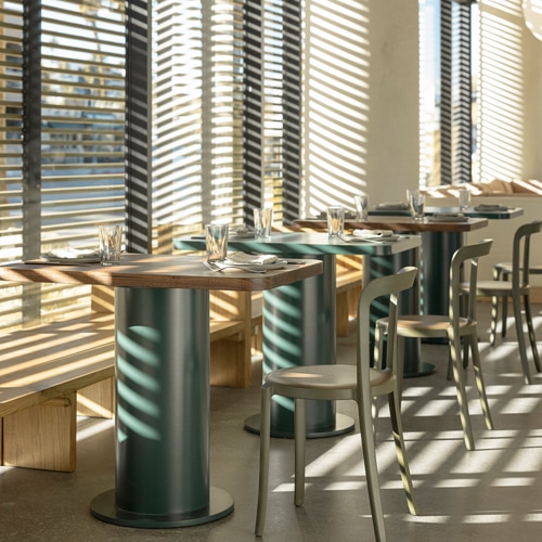 A modern restaurant interior with natural light streaming in through venetian blinds, casting shadows on wooden tables set with glasses and napkins, paired with sleek metal and wood chairs.